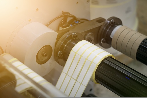 A machine for the production of white self-adhesive labels. The shafts of the machine for winding rolls of tape with a label. Die-cutting and cutting of paper for label production. Selective focus.